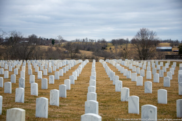 A central aisle runs between rows of white headstones toward distant trees and a small outbuilding.