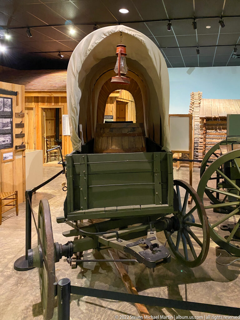 A green wooden wagon with a canvas cover stands in a gallery, viewed from behind between exhibits.