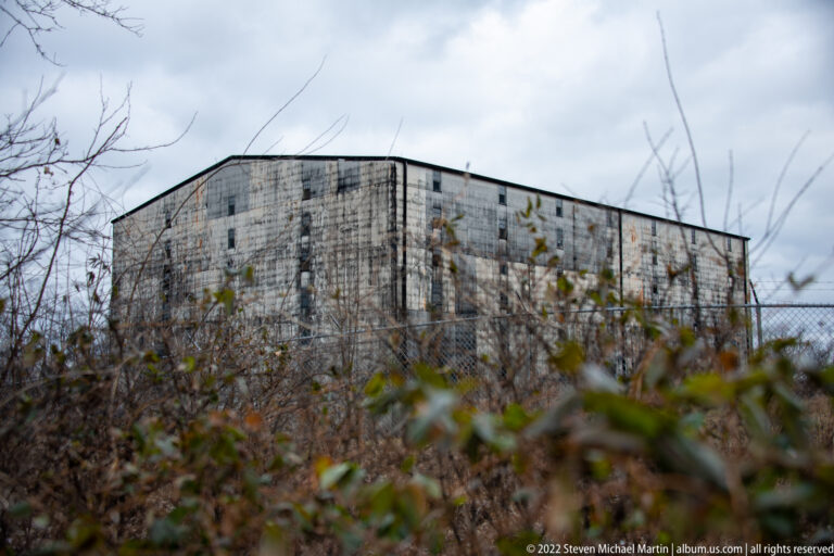 A tall weathered metal-sided building stands behind a chain-link fence and winter brush.