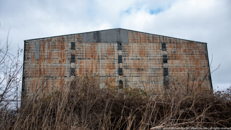 A large corrugated metal building with rust-streaked walls rises behind leafless brush beneath a cloudy sky.