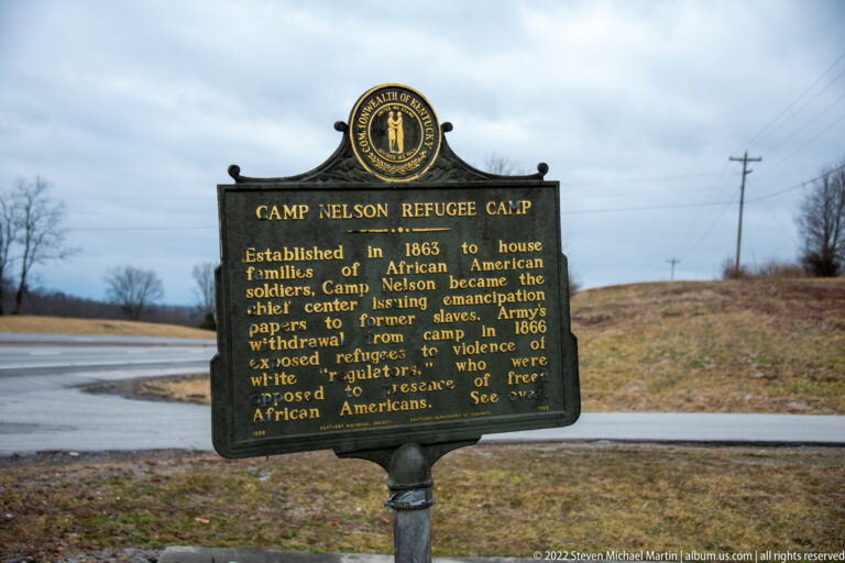 A black historical marker with gold lettering stands beside a road and open winter grass.
