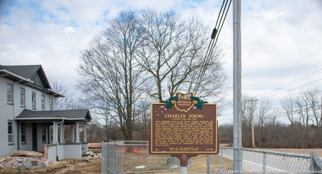 A Charles Young historical marker stands in front of a pale two-story house enclosed by construction fencing.