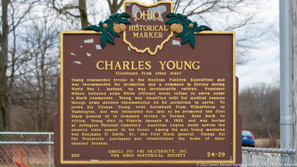 Gold lettering on the reverse side of an Ohio historical marker about Charles Young, with a chain-link fence behind it.