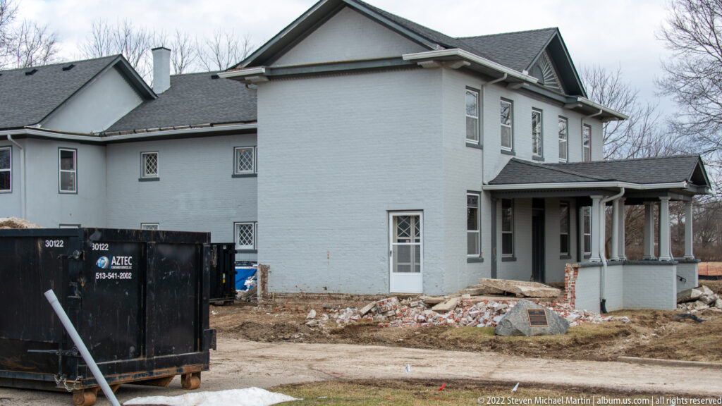 A pale two-story house with a gabled roof stands beside a construction dumpster and scattered masonry debris.