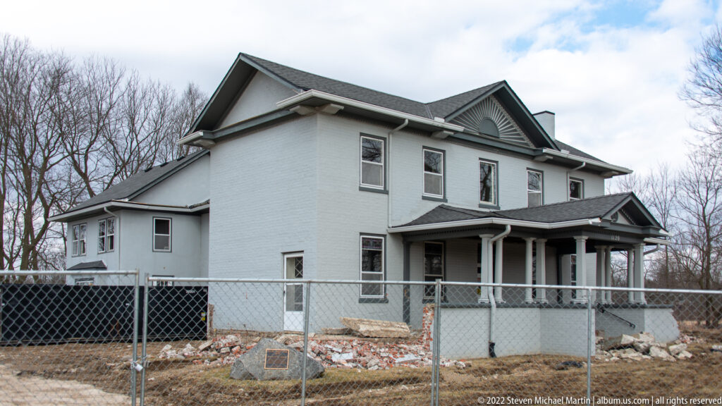 A pale two-story house with a columned porch and layered gables stands behind temporary fencing.