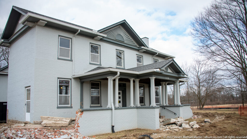 A closer angled view shows the house’s white porch columns, windows, and exposed foundation amid restoration debris.