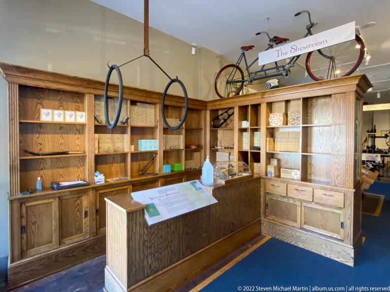 A wooden shop counter and shelves sit beneath a suspended bicycle, hanging rims, and a Showroom sign.