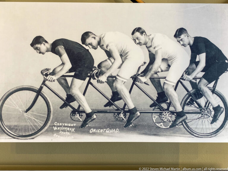 Four men ride an Orient Quad tandem bicycle in a side-view black-and-white photograph.