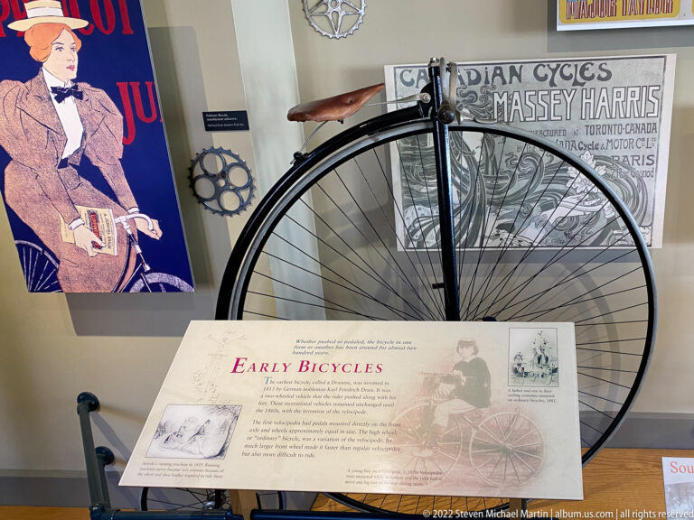 A high-wheel bicycle stands behind an Early Bicycles panel, with a Massey-Harris poster on the wall.