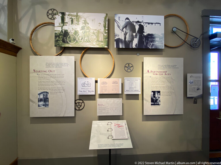 Bicycle wheels, photographs, documents, and interpretive panels fill a museum wall above an open binder.