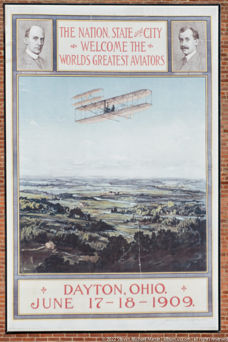 A framed 1909 Dayton poster shows an early aircraft over a landscape beneath a commemorative heading.