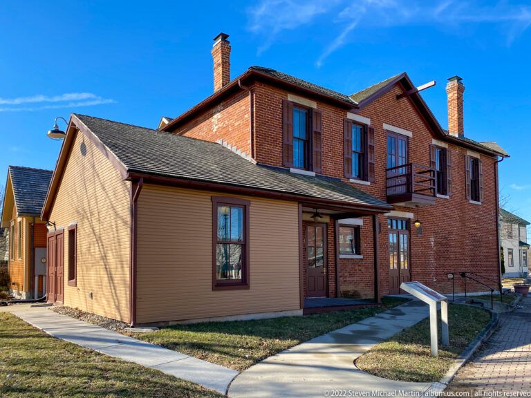 A red-brick upper building rises behind a lower clapboard structure beside a curving walkway and small sign.
