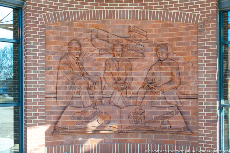 A sculpted wall relief shows three seated men around a table, with an aircraft above and bicycle forms below.
