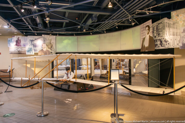 A large early biplane with fabric-covered wings stands behind ropes beneath a curved band of aviation photographs.