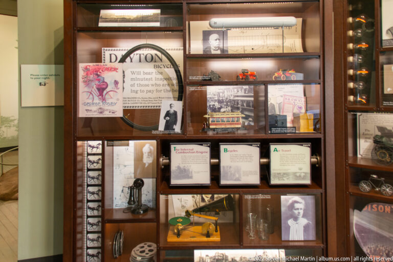 A wooden cabinet displays bicycle parts, advertisements, small models, photographs, and a large wheel.