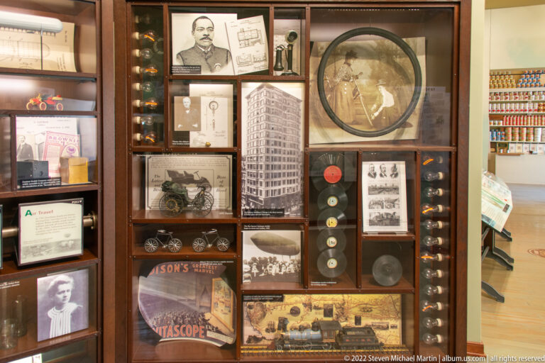 A museum cabinet holds portraits, documents, small mechanical objects, a circular frame, and a model.