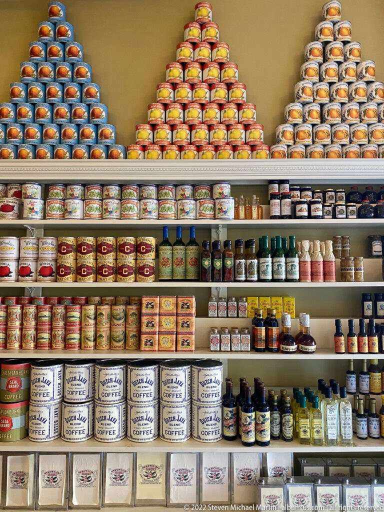 Rows of labeled cans, jars, and bottles fill wooden shelves, with stacked pyramids of cans above.