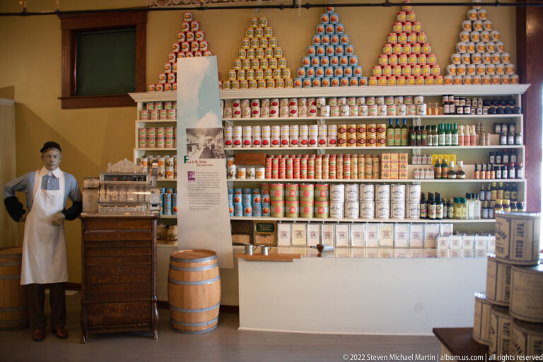 A mannequin shopkeeper stands beside a wooden counter in a recreated store lined with cans, boxes, and jars.