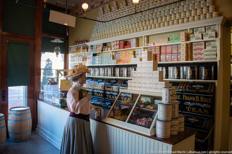 A mannequin in a skirt and brimmed hat stands beside a glass counter beneath shelves of tins and tea boxes.
