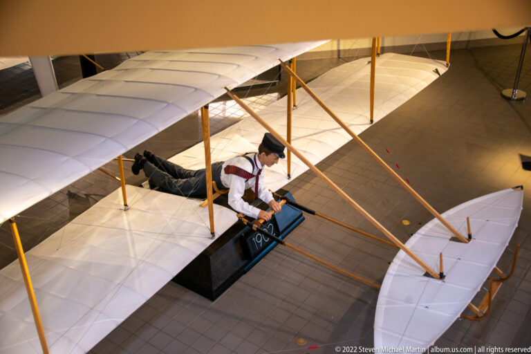 A fabric-wing glider hangs above a prone pilot figure on a central platform, behind visitor barriers.
