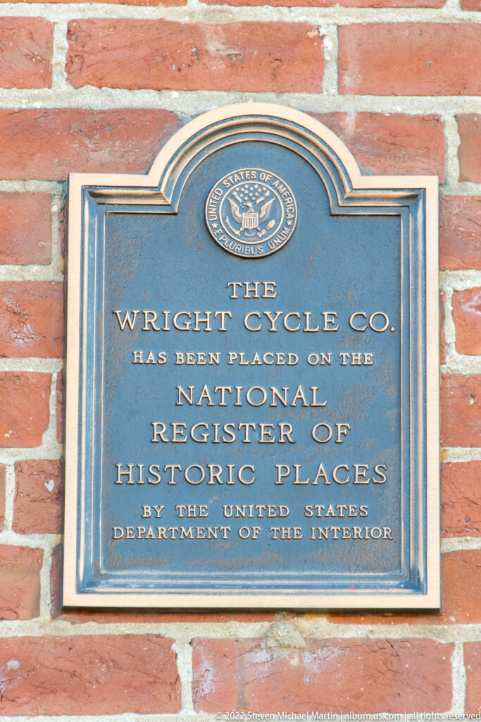 A metal plaque on a brick wall identifies the Wright Cycle Co. as listed on the National Register of Historic Places.