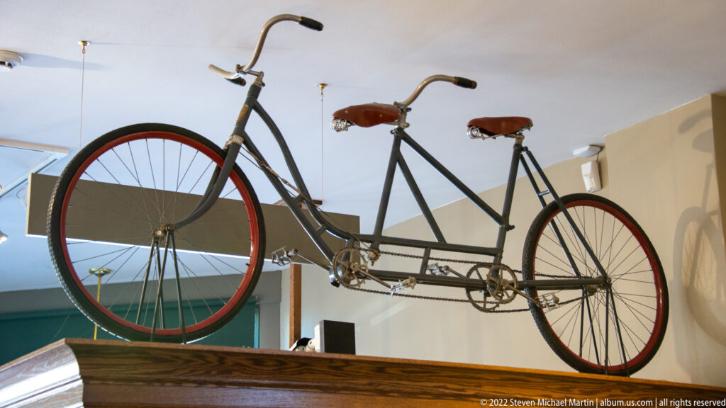 A tandem bicycle with two saddles and two handlebars is displayed above a wooden platform.