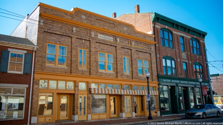 Brick commercial buildings line a Dayton street, with the park sign on a green-trimmed façade at right.