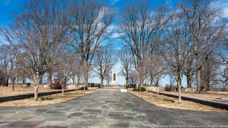 A stone walkway passes between bare trees and low walls toward a slender monument in the distance.