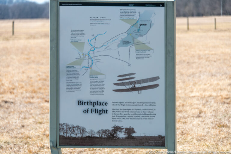A “Birthplace of Flight” wayside panel displays a map, aircraft diagrams, and explanatory text beside an open field.