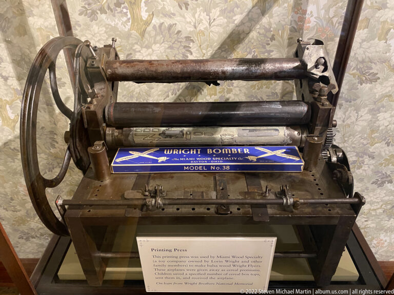 A black iron printing press with a large flywheel and rollers stands above an interpretive label.