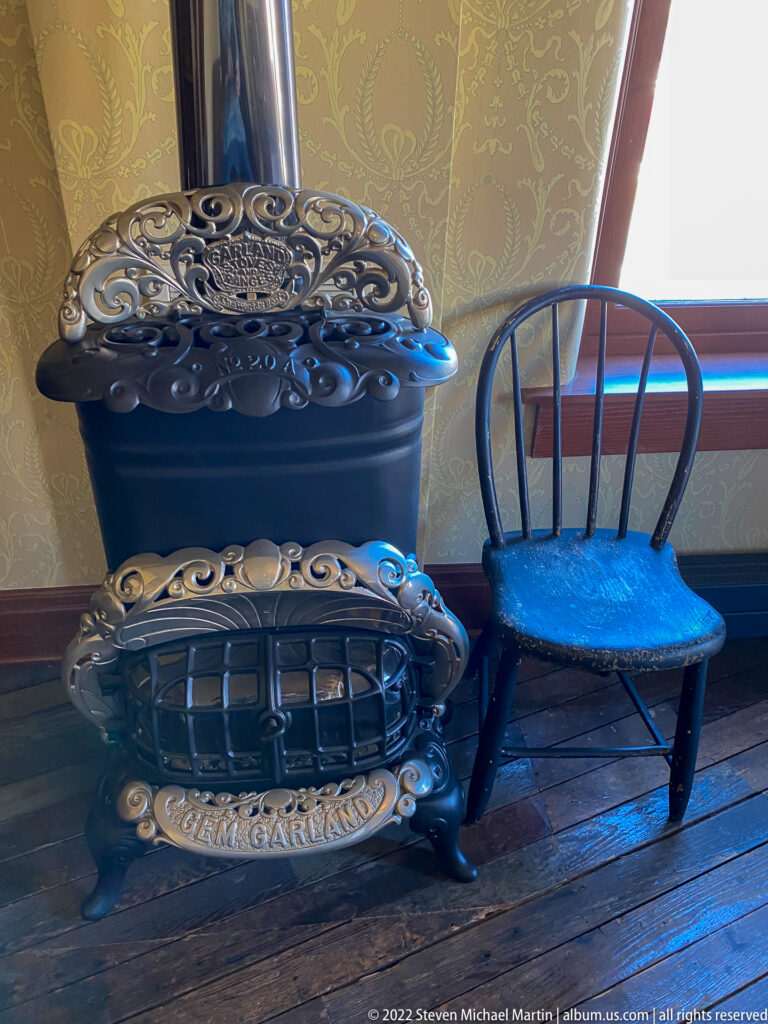 An ornate black cast-iron stove with raised scrollwork stands beside a wooden chair near a window.