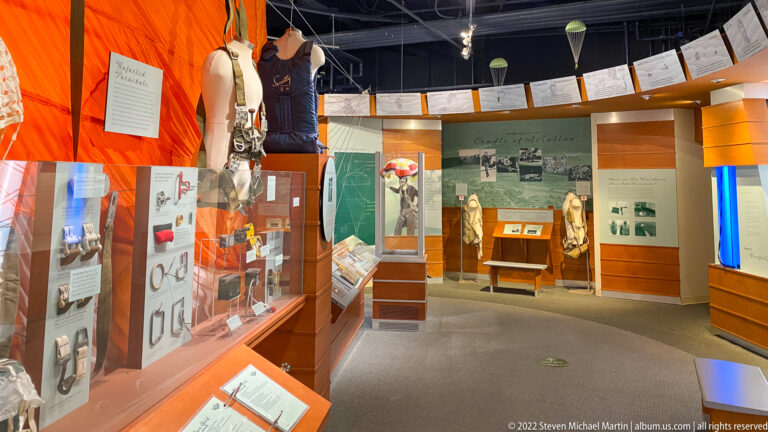 A museum gallery contains orange and green panels, glass cases, aviation mannequins, benches, and hanging aircraft models.