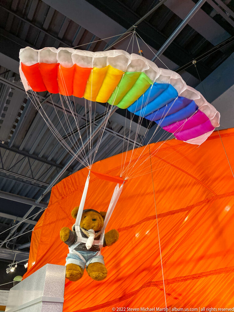 A small teddy bear hangs beneath a rainbow parachute in front of a large orange canopy.