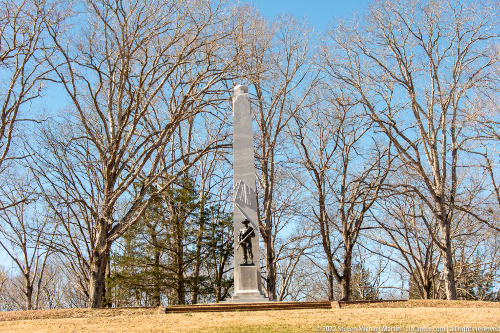 A pale stone obelisk with a small figure near its base stands in a leafless wooded clearing.