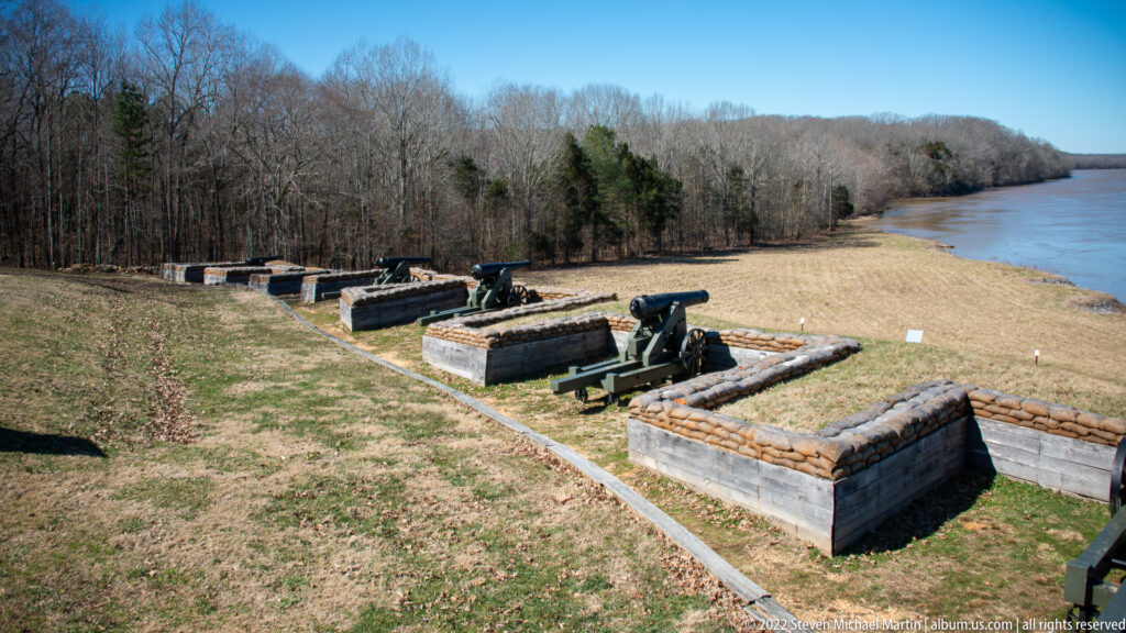 Several cannon emplacements line a grassy slope above a river, with low timber and earth walls around each gun.
