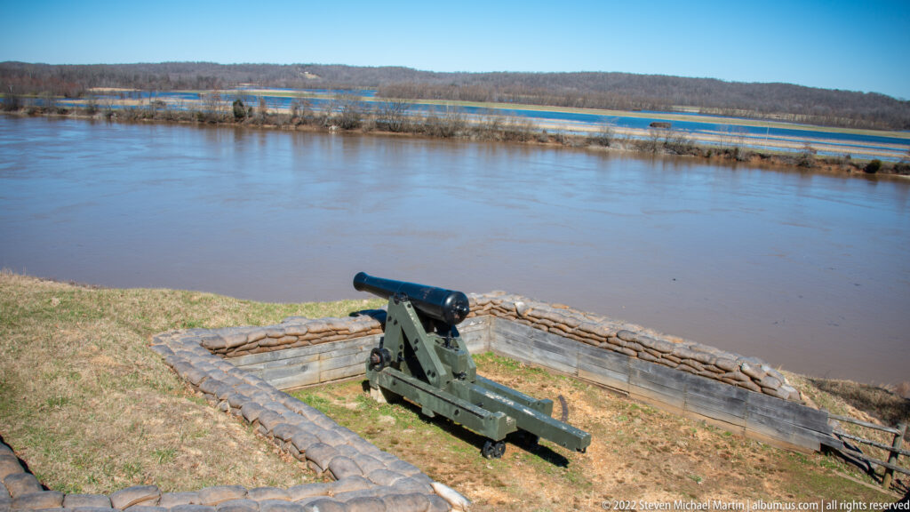 A green cannon rests in a low timber-and-earth emplacement overlooking a wide river.