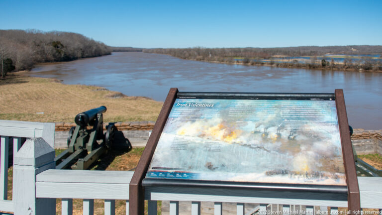 A cannon and slanted wayside panel stand at a wooden-railed overlook above a broad river.