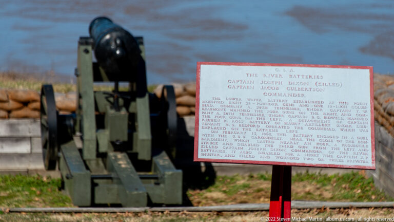 A dark cannon and green carriage stand beside a white river-batteries sign overlooking water.