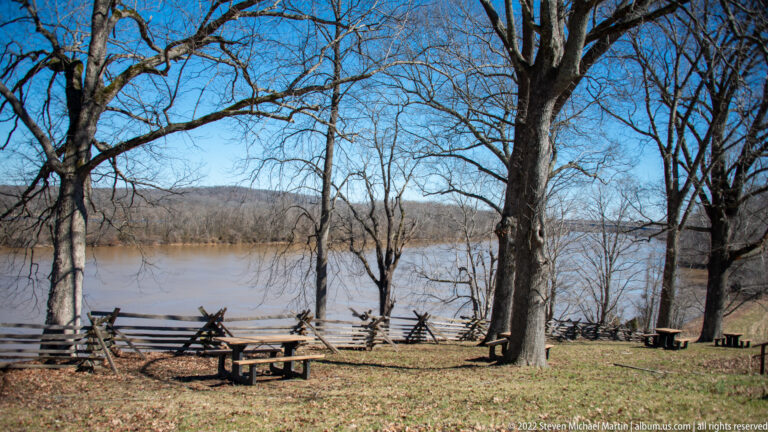 A split-rail fence and picnic tables sit beneath bare trees beside the river.
