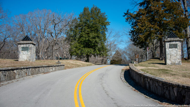 A paved road curves between stone walls and pillars topped with cannonballs at a park entrance.