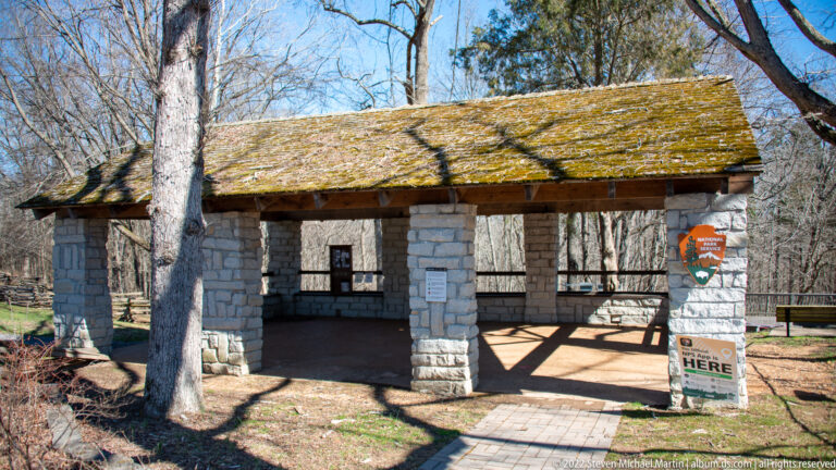 A stone-column pavilion with a mossy green roof stands among trees beside a “You Are Here” sign.