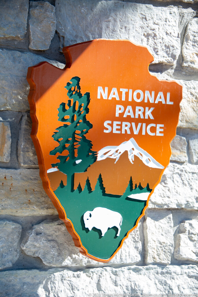 An orange National Park Service arrowhead with a tree, mountain, and bison is mounted on a pale stone wall.