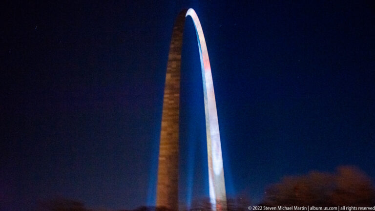 The illuminated Gateway Arch rises in a narrow curve against a deep blue night sky.