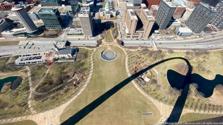 The arch casts a long curved shadow across lawns, paths, and reflecting pools.
