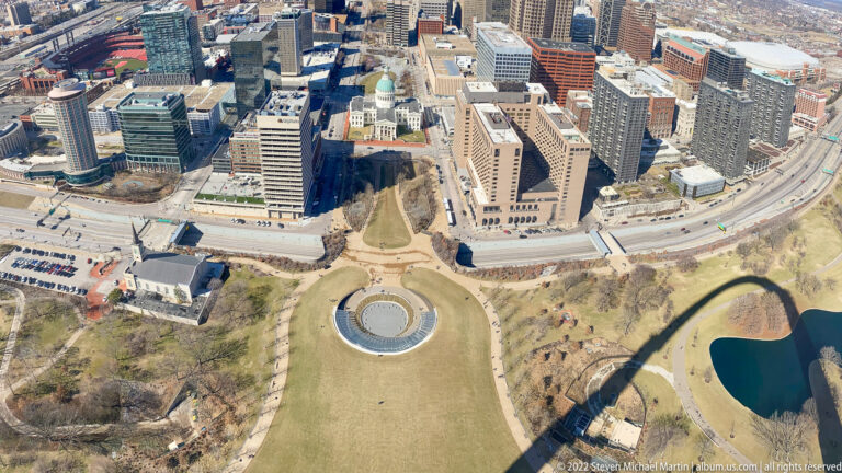 An aerial view shows the park lawn and circular pool below downtown, crossed by the arch’s shadow.