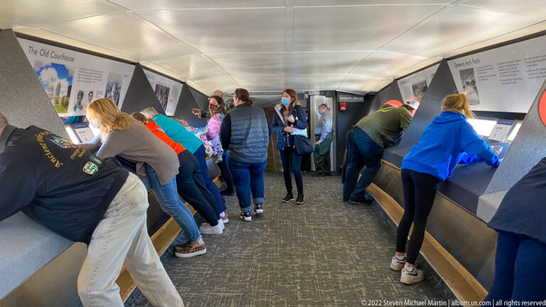 Visitors lean over exhibit stations along both sides of a narrow museum gallery.