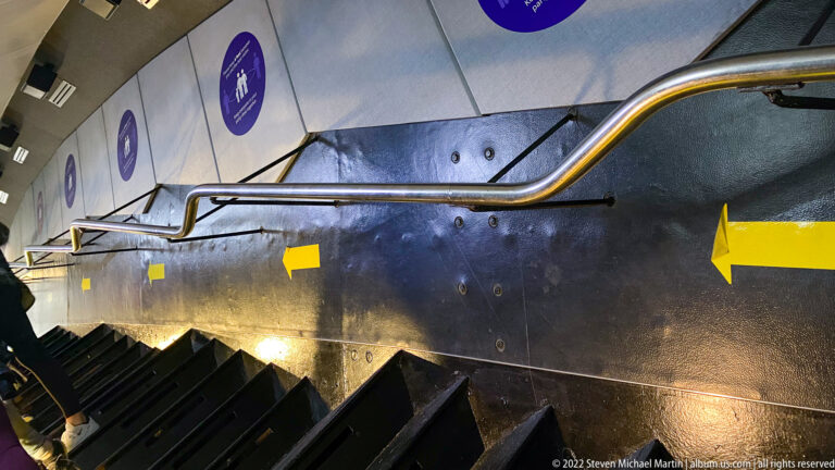 A steep dark stairway descends beneath metal handrails and yellow directional arrows.