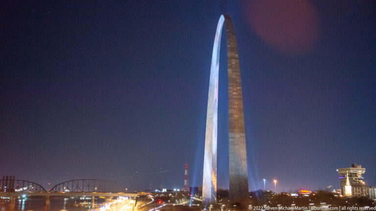 The lit Gateway Arch stands above riverfront roads, with a bridge and city lights at left.