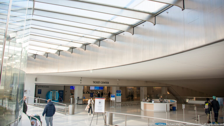 A bright visitor-center hall has a glass roof, ticket counters, and visitors crossing the floor.
