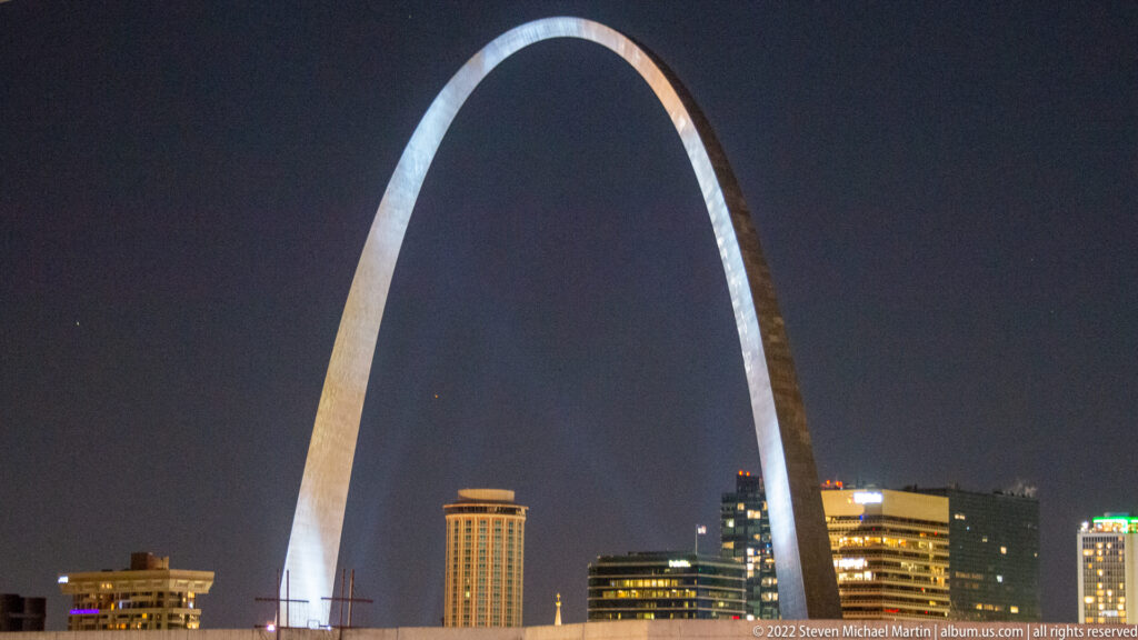 The Gateway Arch frames illuminated downtown buildings beneath its curved crown at night.